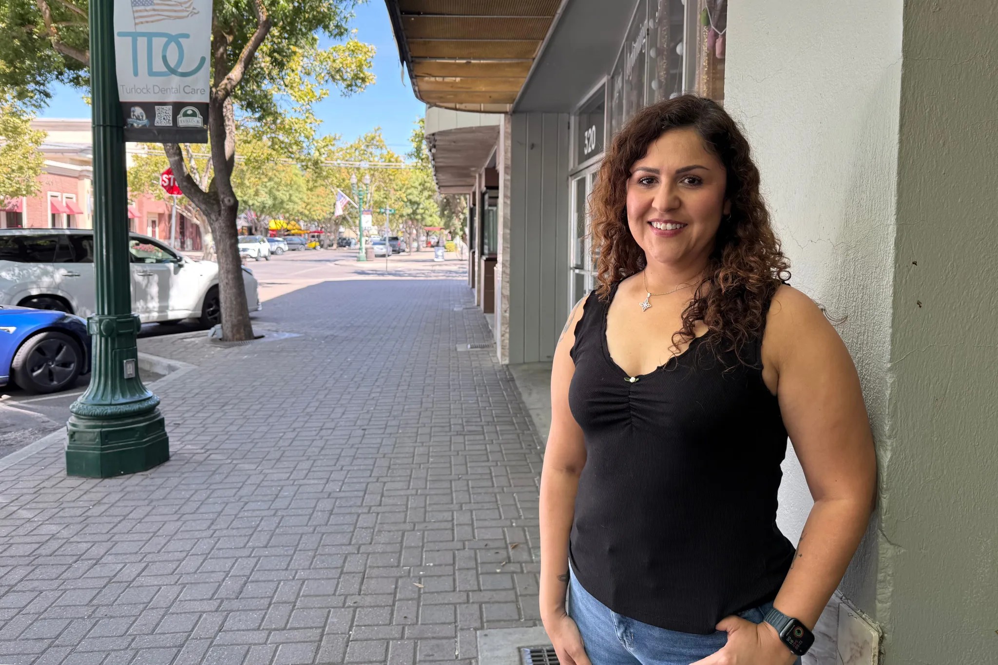 Person standing on a sidewalk, smiling, with trees and cars in the background.